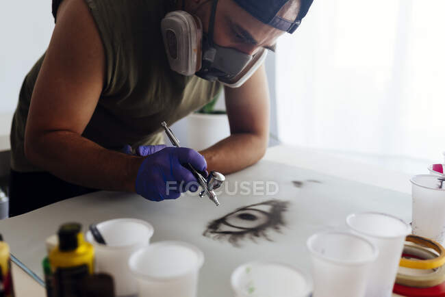 Close up of airbrush painter with mask drawing an eye on a paper. — Stock Photo