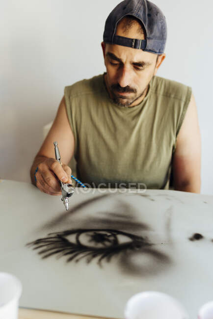 Close up of airbrush painter with cap drawing an eye on a paper. — Stock Photo