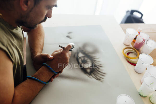 Aerial view of airbrush painter with cap drawing an eye on a paper. — Stock Photo