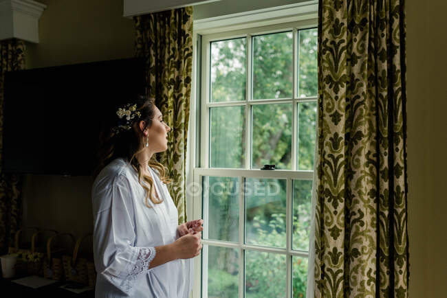 Una novia preparándose mirando por la ventana de su habitación de hotel - foto de stock
