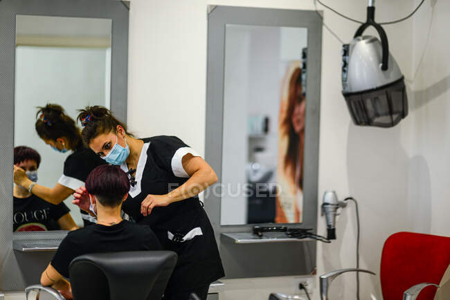 Estilista de cabelo feminino no trabalho usando máscara facial enquanto estiliza jovem — Fotografia de Stock
