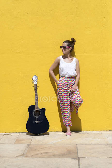 Guitarrista bonita mujer mirando hacia otro lado con la guitarra apoyada en la pared amarilla - foto de stock