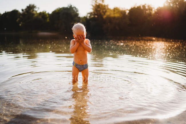 Joven niño blanco cubriendo la cara de pie en el agua en el lago luz dorada - foto de stock