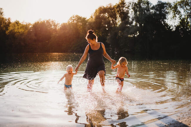Madre e hijos pequeños caminando fuera del lago tomados de la mano sonriendo - foto de stock