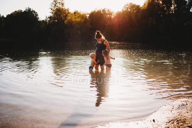 Bambini che baciano la pancia della madre incinta in acqua al lago durante il tramonto — Foto stock