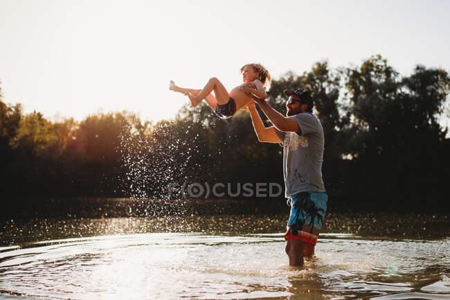 Отец держит маленького ребенка в воздухе у озера, брызгая водой — стоковое фото