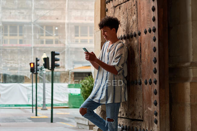 Young Afro apoyado en una puerta de madera mirando su teléfono - foto de stock