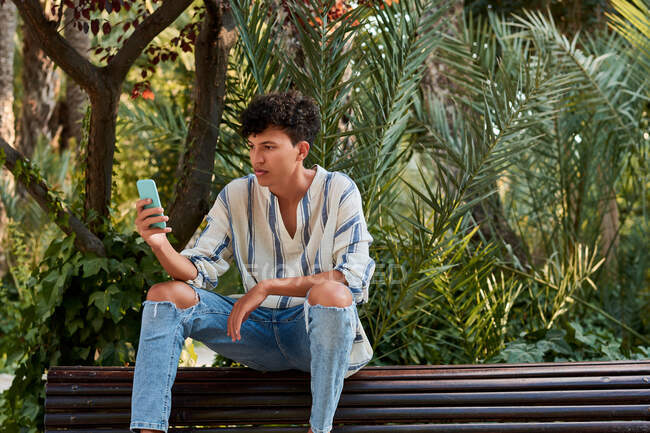 Молодой человек с афроволосами наблюдает за своим смартфоном, сидящим на — стоковое фото