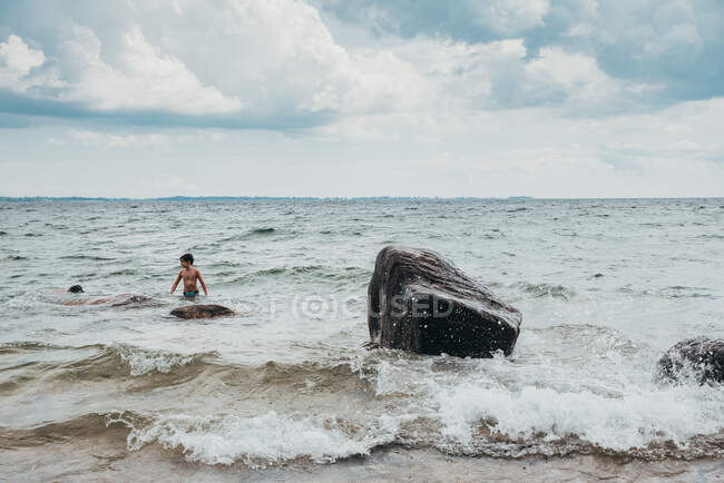 Young boy sitting on big rock in lake getting splashed on summer day. — Stock Photo
