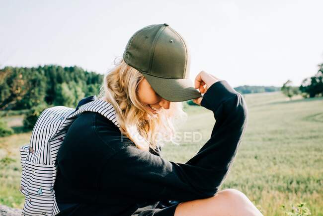 Woman sat in the countryside holding her cap over her face looking shy — Stock Photo