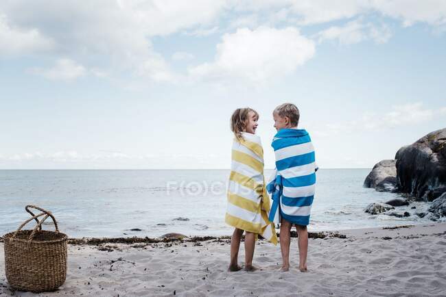 Hermano y hermana se pararon riendo en la playa envueltos en toallas - foto de stock