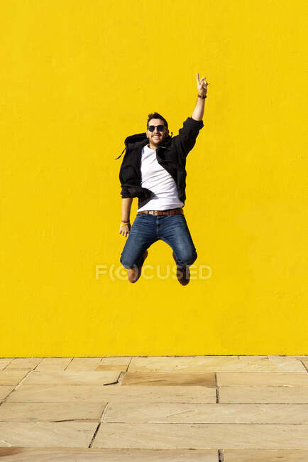 Joven con gafas de sol saltando frente a una pared amarilla. - foto de stock