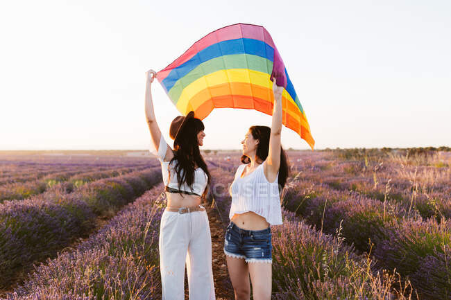 Namoradas olhando umas às outras em um campo de lavanda acenando uma bandeira lgbt — Fotografia de Stock