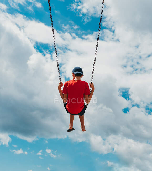 Vue arrière du garçon sur une balançoire avec seulement ciel nuageux en arrière-plan. — Photo de stock