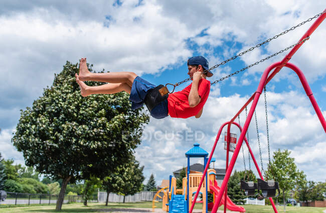 Vista lateral do menino em um balanço com parque infantil no fundo. — Fotografia de Stock