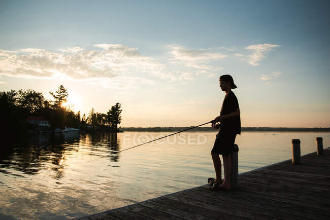 Adolescent boy fishing off dock on lake at sunset in Ontario, Canada. — Stock Photo