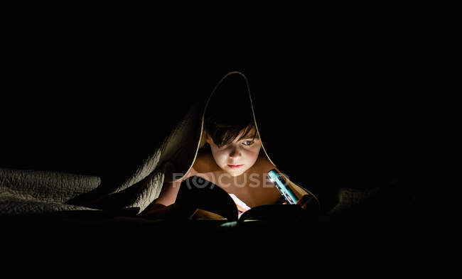 Молодий хлопчик читає книгу під ковдрою, використовуючи ліхтарик . — стокове фото
