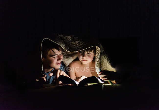 Hermanos leyendo un libro bajo una manta por la linterna en la noche. - foto de stock