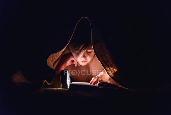 Young boy reading a book under a blanket using a flashlight. — Stock Photo