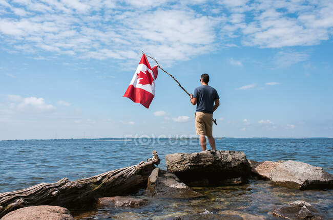 Mann mit Kanada-Fahne am felsigen Ufer eines Sees an einem Sommertag. — Stockfoto