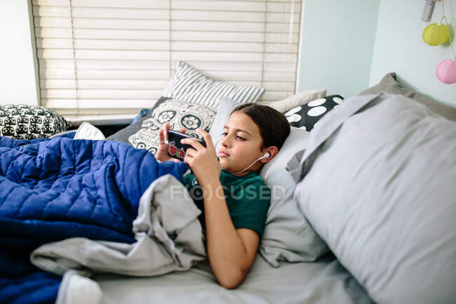Zwillingsmädchen liegt mit Kopfhörern im Bett und schaut auf ihr Handy — Stockfoto