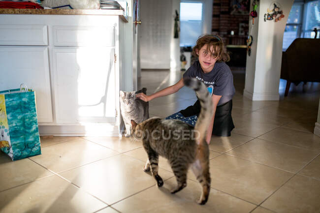 Teen girl sits on the tile floor petting one of two cats — Stock Photo