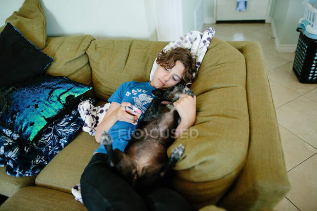 Дівчина лежить на дивані, дивлячись на телефон і лоскочучи свого маленького собаку — стокове фото