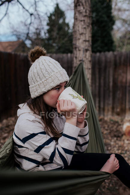 Девушка-подросток, сидящая в гамаке и пьющая из кружки — стоковое фото
