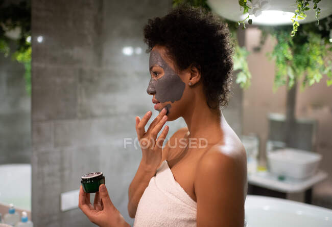 Vista laterale della femmina nera che applica maschera di fango sul viso durante la routine di cura della pelle a casa — Foto stock