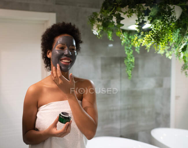 Feliz adulto preto fêmea sorrindo e aplicando máscara de barro no banheiro em casa — Fotografia de Stock