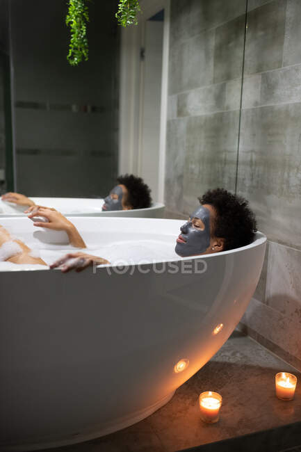 Erwachsene afrikanisch-amerikanische Frau mit Lehmmaske entspannt sich in der Badewanne während der Wellness-Behandlung zu Hause — Stockfoto