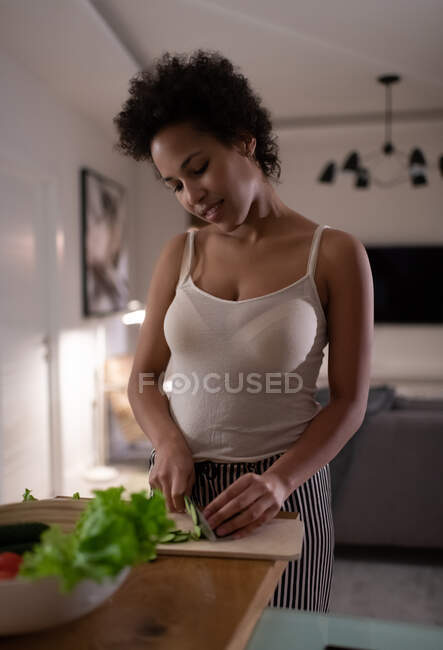 Feliz senhora negra sorrindo e cortando pepino enquanto cozinha salada saudável em casa — Fotografia de Stock
