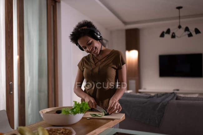 Happy african american lady listening to music and cooking healthy salad for dinner at home — Stock Photo