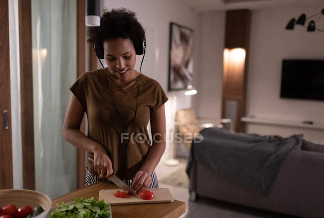 Fröhliche Afroamerikanerinnen hören Musik und schneiden Tomaten für das Abendessen zu Hause — Stockfoto