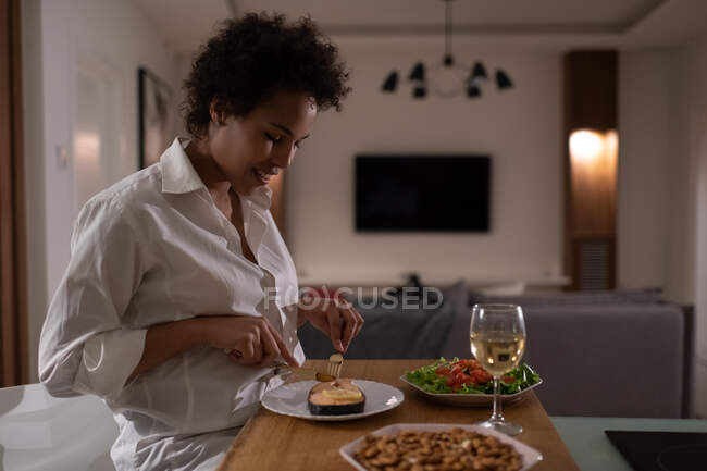 Glad ethnic woman cutting fish while having organic food for dinner at home — Stock Photo