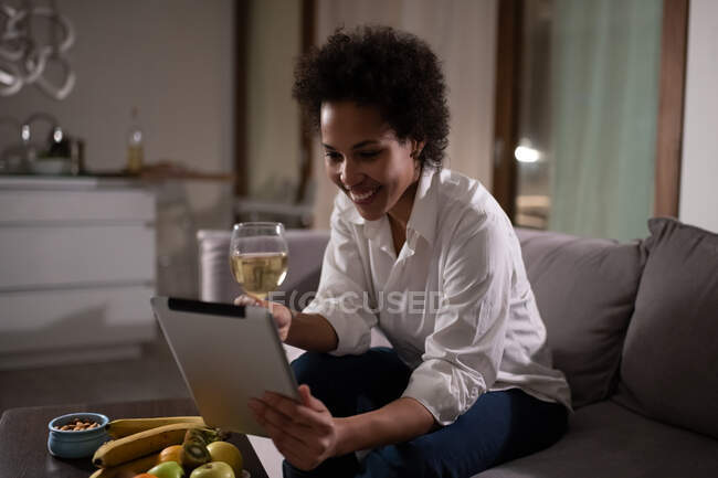 Heureuse dame ethnique avec du vin en utilisant une tablette pour parler avec son petit ami pendant la date en ligne — Photo de stock
