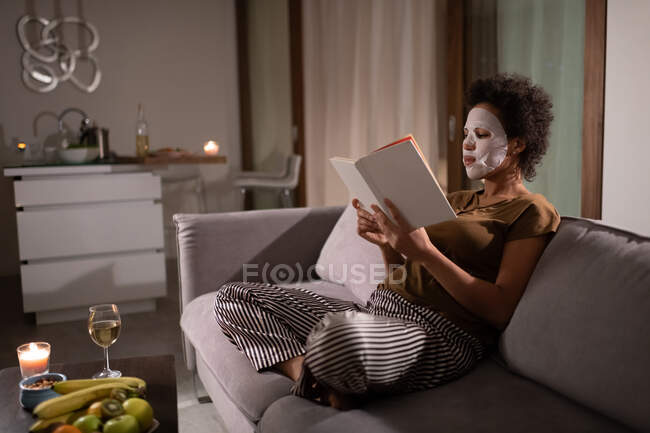 Black female in sheet mask sitting on sofa and reading book during skin care routine — Stock Photo