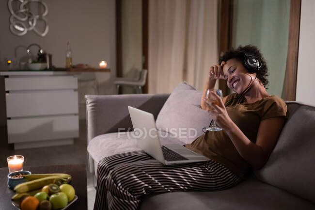Merry ethnic female with wine smiling and watching film on laptop on sofa at home — Stock Photo