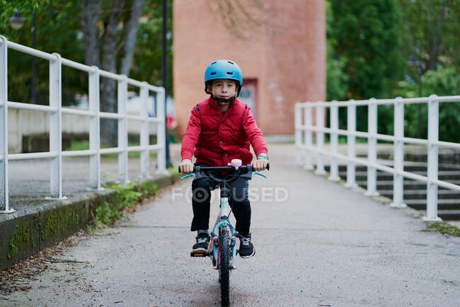 Menino andando de bicicleta com um capacete azul e um sobretudo vermelho — Fotografia de Stock
