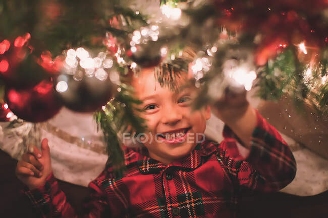 Boy hanging looking at camera under the Christmas Tree at night time — Stock Photo