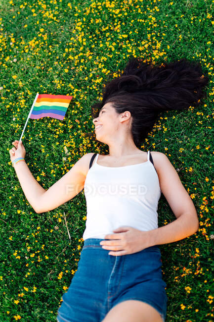 Glückliche Frau liegt mit Blumen im Gras und schwenkt eine lgtb-Fahne — Stockfoto