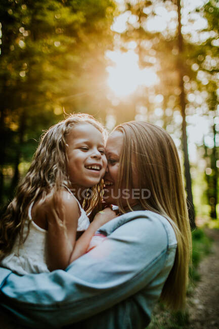 Feliz mamá e hija riendo y acurrucándose en el bosque retroiluminado - foto de stock