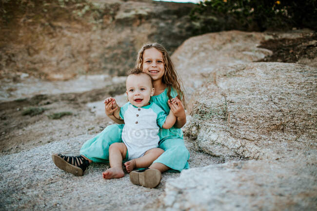 Felice giovane ragazza seduta e tenendo il fratellino su una grande roccia — Foto stock