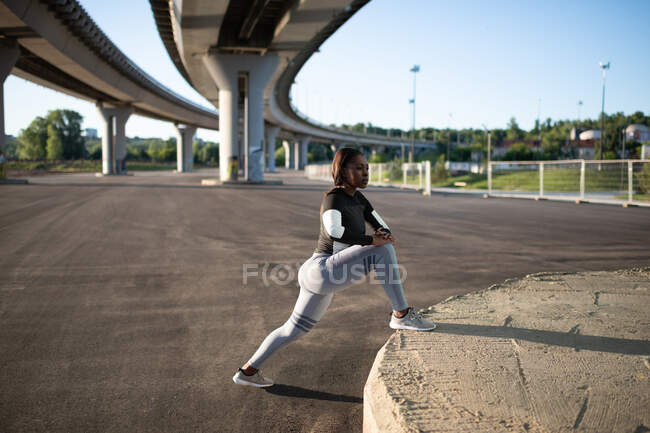Side view of black female athlete lunging under bridge in morning in city — Stock Photo