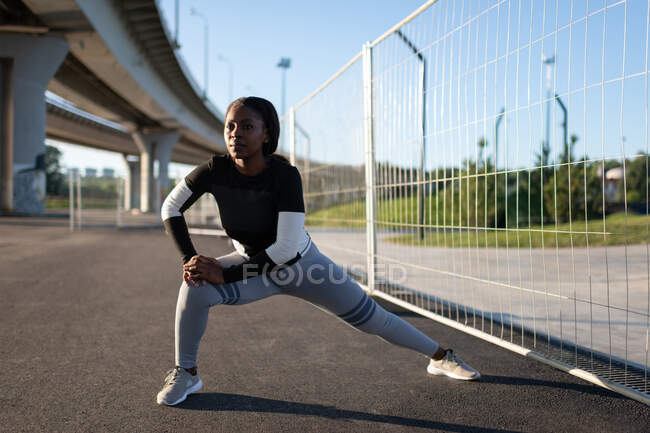 Atleta feminina negra pulando perto da cerca enquanto se aquece de manhã na cidade — Fotografia de Stock