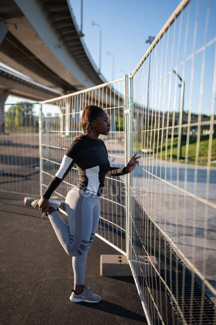 Seitenansicht der ethnischen Läufer Stretching Bein in der Nähe von Zaun während des morgendlichen Trainings in der Stadt — Stockfoto