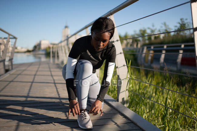 Ethnic female athlete tying shoelaces during running workout on quay in morning — Stock Photo