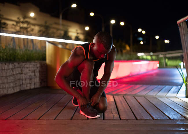 Deportista negro atando cordones antes de correr entrenando de noche en la ciudad - foto de stock