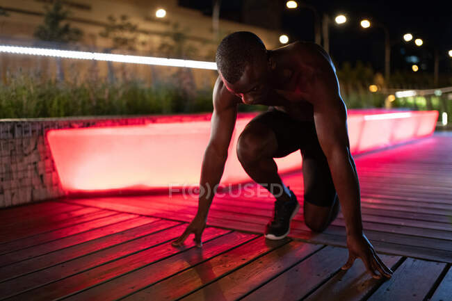 Deportista étnico sin camisa en posición de salida agachado listo para correr por la noche en la ciudad - foto de stock