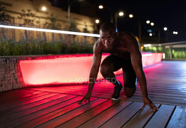 Atleta étnico listo para correr en el camino durante el entrenamiento de fitness por la noche en la ciudad - foto de stock